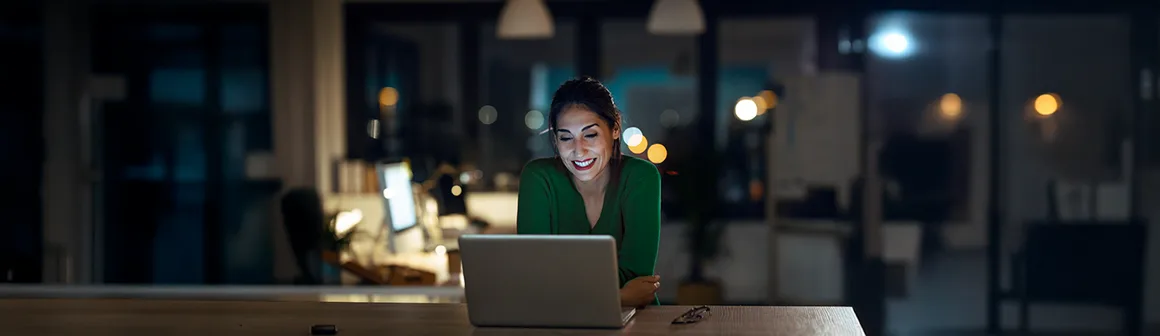 Mulher sorrindo, sentada em uma mesa, com um notebook aberto, em uma sala a meia luz, aprendendo como fazer assinatura digital