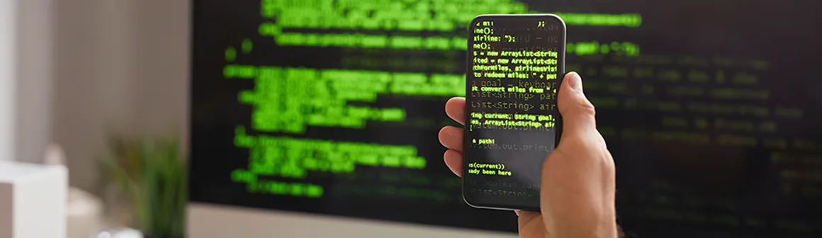 Mão segura um smartphone exibindo linhas de código verde, com uma tela de monitor ao fundo, representando o desenvolvimento de inteligência artificial.
