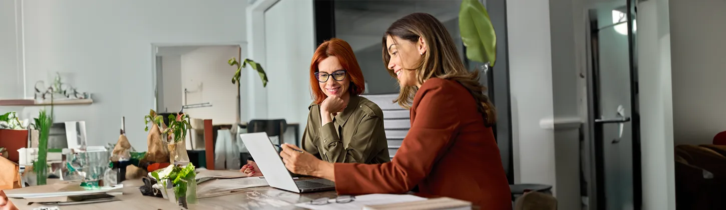 Duas mulheres estão sentadas lado a lado em uma mesa, olhando atentamente para a tela de um laptop aberto. A cena sugere uma reunião de trabalho ou uma colaboração em um projeto, possivelmente envolvendo a análise de documentos ou informações para uma ação revisional, dado o foco e a concentração das participantes. A mesa está organizada com papéis, plantas e outros objetos de escritório, criando um ambiente de trabalho profissional e criativo.