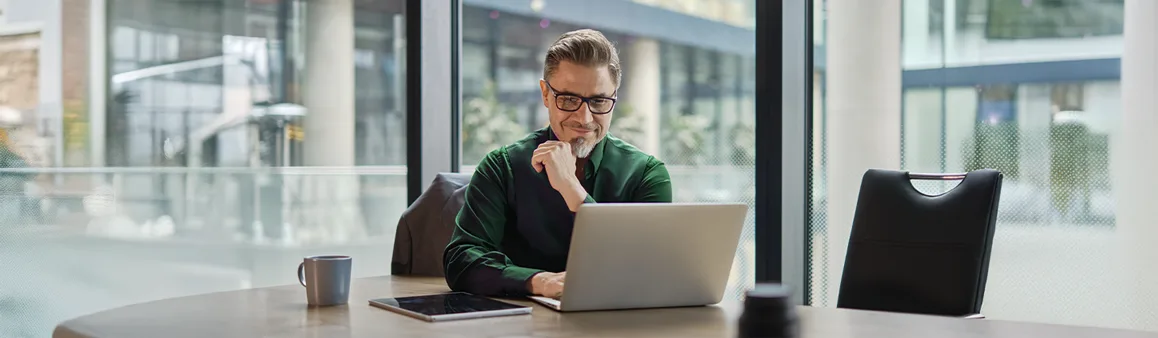 Um homem de óculos, vestindo uma camisa verde, está concentrado em seu laptop em um escritório moderno com grandes janelas. Ele tem uma das mãos no queixo, parecendo pensativo. A cena sugere um ambiente de trabalho focado em tecnologia e inovação, possivelmente explorando conceitos como blockchain ou outras soluções digitais avançadas. Um tablet e uma caneca estão na mesa ao seu lado.