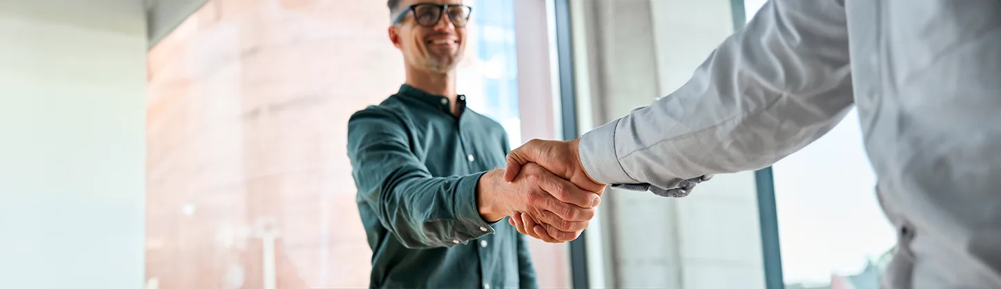 Dois homens em ambiente de escritório apertam as mãos, sorrindo. O aperto de mãos pode simbolizar um acordo de negócios ou uma parceria, representando a união de capital social e recursos para um objetivo em comum.