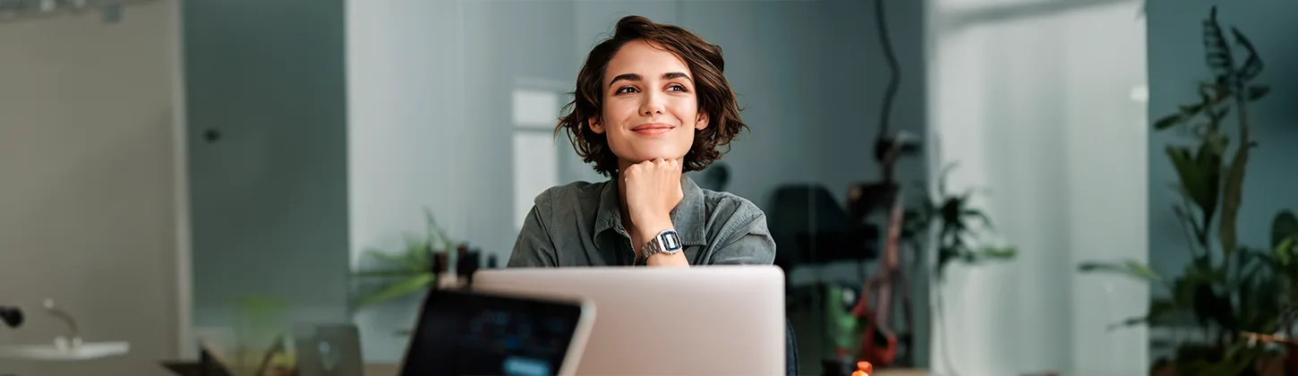 Uma mulher com cabelo curto e escuro, usando uma camisa verde e um relógio, sorri pensativamente enquanto olha para cima e para a direita, com as mãos cruzadas sob o queixo, em frente a um laptop. A cena pode sugerir que ela está refletindo sobre questões financeiras ou de planejamento patrimonial, como o imposto de herança.
