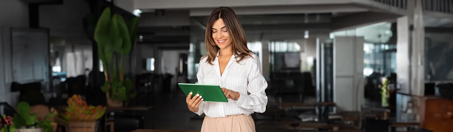 Uma mulher sorridente, vestindo uma camisa branca listrada e calças bege, está olhando para um tablet verde em suas mãos. Ela parece estar em um ambiente de escritório ou um espaço de trabalho moderno, possivelmente revisando ordens de compra ou informações relacionadas a negócios. O fundo está ligeiramente desfocado, sugerindo foco na mulher e no dispositivo.