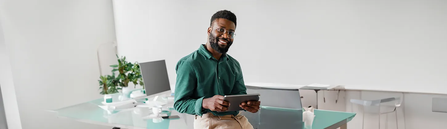 Um homem sorridente, com barba e óculos, veste uma camisa verde-escura e calças claras, segurando um tablet. Ele está em um escritório moderno com plantas e monitores, demonstrando confiança, como se estivesse pronto para implementar um plano de ação.