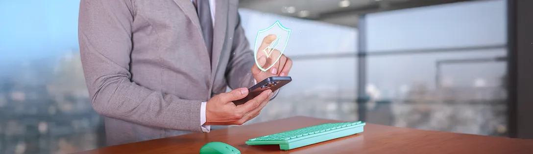 Um homem em um terno cinza e gravata está segurando um smartphone, com um ícone de escudo e um tique de verificação verde brilhando acima dele, simbolizando segurança digital. A imagem sugere a importância da proteção de dados e autenticação, como a utilizada em sistemas de SSO (Single Sign-On). Um teclado verde e um mouse também estão visíveis na mesa de madeira.