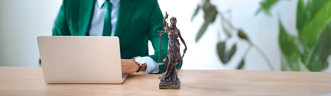 Um profissional, vestido com um terno verde e gravata, está sentado em uma mesa de madeira com um laptop à frente. Ao lado, uma estátua da Justiça (Têmis) com olhos vendados e balança, simbolizando o direito e a lei. A cena sugere um ambiente legal ou jurídico, onde a necessidade de verificar assinatura digital em documentos é crucial para a autenticidade e validade.