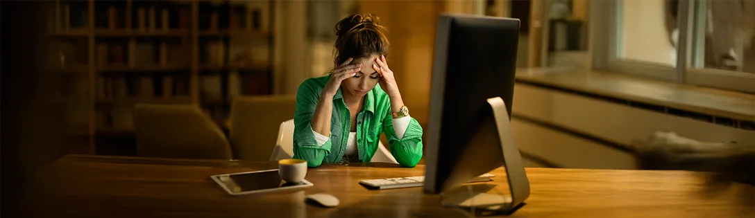 Uma mulher com as mãos na cabeça parece estressada diante do computador, possivelmente tentando entender o que é IOF e suas implicações financeiras.