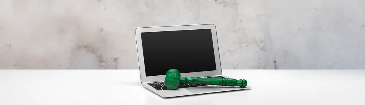 Um notebook prateado com a tela preta está sobre uma superfície branca, e um martelo de juiz verde-esmeralda repousa sobre o teclado. O martelo, símbolo da justiça e da lei, em conjunto com o laptop, evoca a discussão sobre a regulamentação da inteligência artificial e suas implicações legais. O fundo é uma parede de concreto com tons acinzentados e brancos.