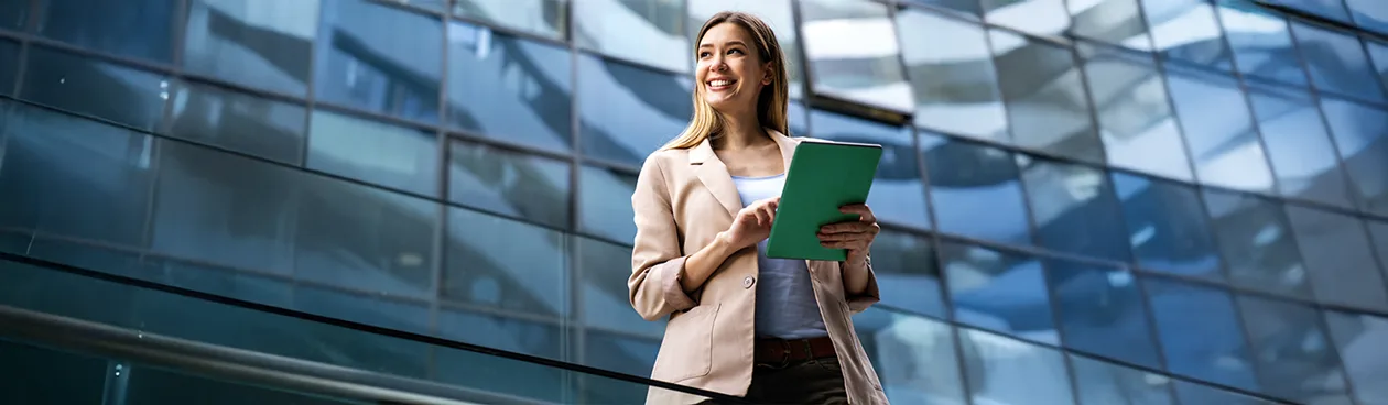 Uma mulher sorridente e bem-vestida segura um tablet verde em frente a um edifício moderno com fachada de vidro, refletindo o céu. A imagem evoca o dinamismo do mundo corporativo e tecnológico, associado ao ambiente das Big Techs.