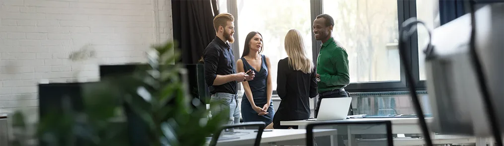 Quatro colegas de trabalho conversam em um escritório moderno, com grandes janelas e paredes de tijolo branco. A interação descontraída sugere flexibilidade no ambiente de trabalho, o que pode estar alinhado com o conceito de chronoworking, que busca otimizar a produtividade ao respeitar os ritmos biológicos de cada indivíduo.