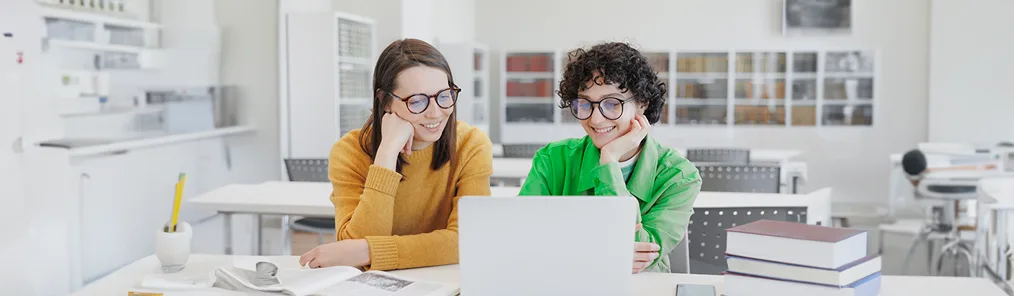 Duas mulheres jovens e sorridentes, ambas usando óculos, estão sentadas em uma mesa em um ambiente de escritório ou sala de aula, olhando para um laptop à frente delas. Elas parecem estar colaborando ou estudando juntas, possivelmente explorando conceitos tecnológicos complexos como o de smart contract. Uma delas veste um suéter mostarda e a outra um casaco verde, e há livros e papéis na mesa.