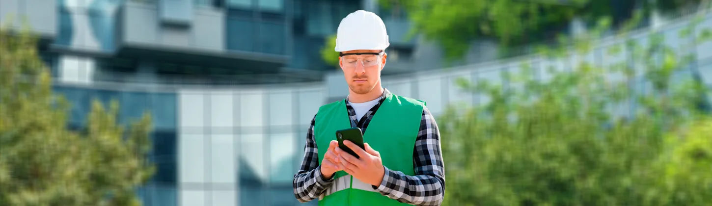 Um homem com capacete branco e colete verde em um canteiro de obras está usando um smartphone, o que pode representar o uso da assinatura eletrônica e digital na construção civil para agilizar processos, como no case Cyrela.