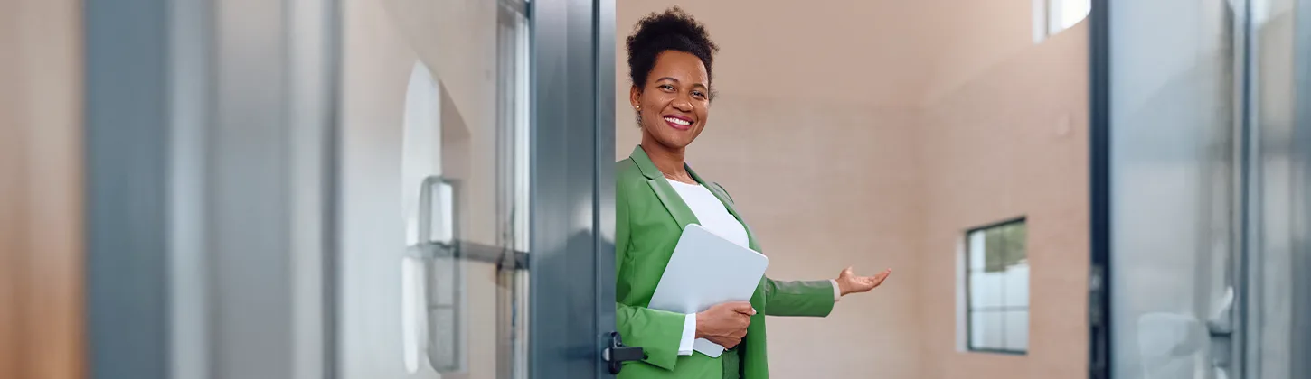 Uma mulher sorridente com blazer verde e tablet na mão abre a porta de uma casa, convidando a entrada. A cena pode representar o processo de usar um checklist de vistoria de imóveis para documentar as condições da propriedade.
