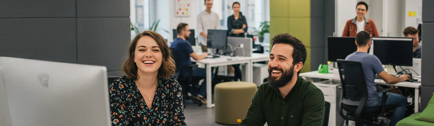 Dois colegas de trabalho sorriem em frente a um computador, enquanto outros colaboradores interagem ao fundo em um escritório movimentado. A imagem transmite um ambiente de trabalho colaborativo e positivo, onde a comunicação interna é eficiente e eficaz.