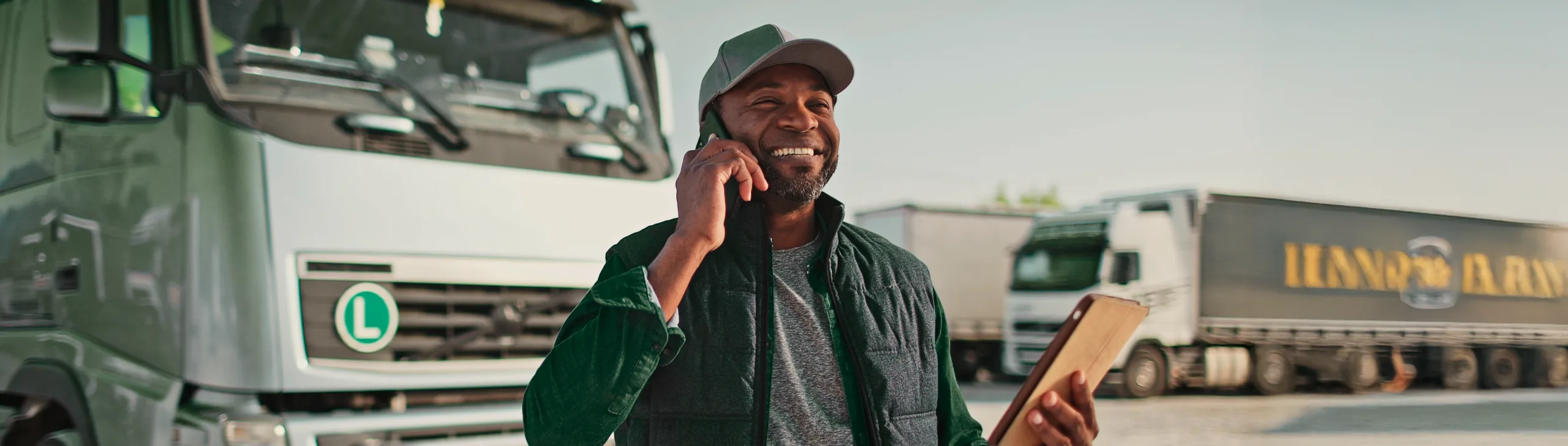 Um motorista de caminhão sorridente, segurando um celular e uma prancheta, está em um pátio de caminhões. A imagem sugere que ele está finalizando os detalhes de um contrato de frete, destacando a importância da comunicação e da documentação no setor de transporte.