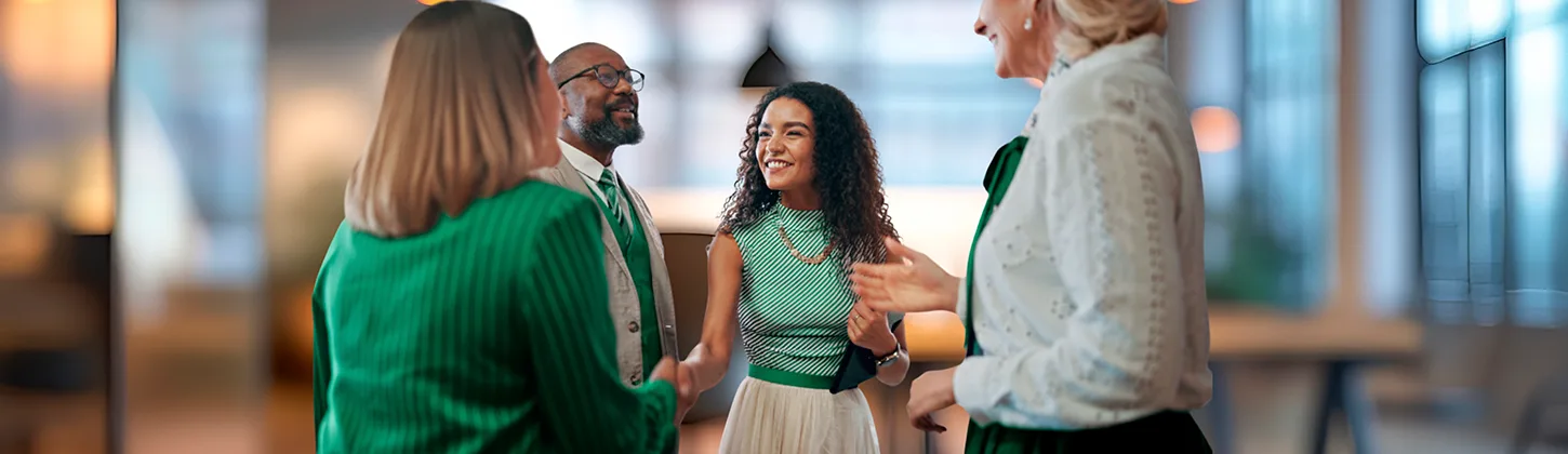 Uma mulher sorridente com uma blusa listrada aperta a mão de outra pessoa em um ambiente de escritório. Outros dois colegas de trabalho sorriem ao redor. A cena simboliza o início ou a finalização de uma Parceria estratégica entre os envolvidos.
