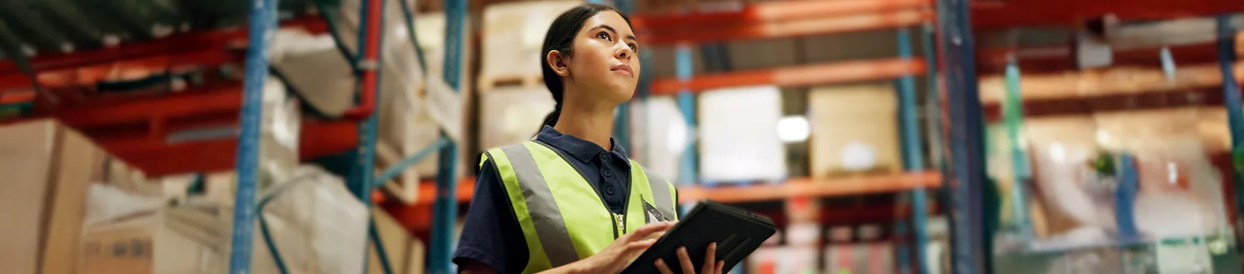 Uma mulher em colete de segurança e uniforme, segurando um tablet, está em um grande armazém com prateleiras cheias. Ela parece estar gerenciando o inventário ou a logística, destacando o uso da assinatura eletrônica para suprimentos e compras para agilizar a documentação de pedidos e recebimentos.