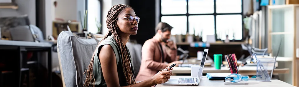 Uma mulher com tranças e óculos, segurando um smartphone, está concentrada em seu trabalho em frente a um laptop em um escritório moderno. A imagem sugere foco e análise de dados, essenciais para otimizar e gerenciar o ciclo de vendas.