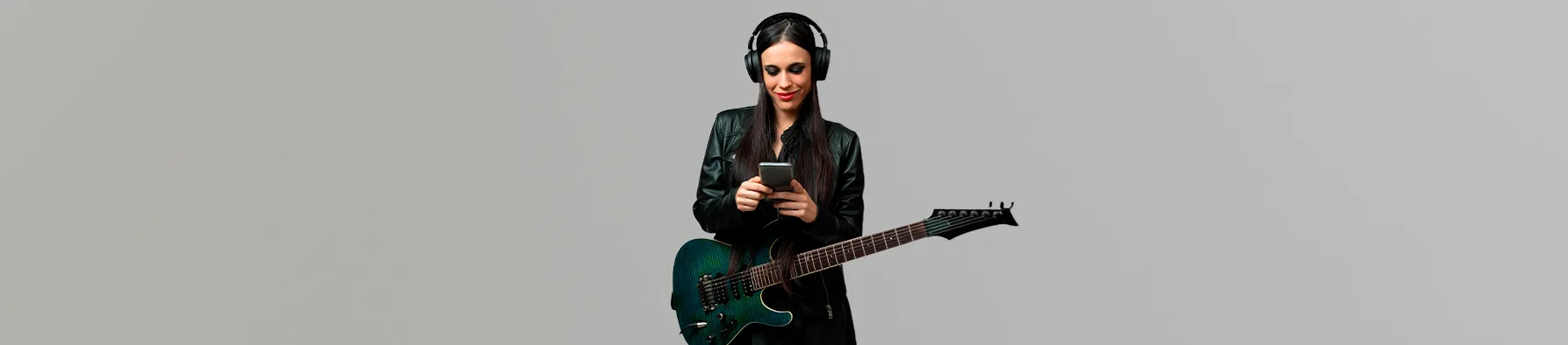 Uma mulher sorridente com fones de ouvido e uma jaqueta de couro preta segura uma guitarra elétrica verde escura e um celular. A imagem representa músicos na era digital, sugerindo a facilidade de gerenciar contratos para músicos eletronicamente.
