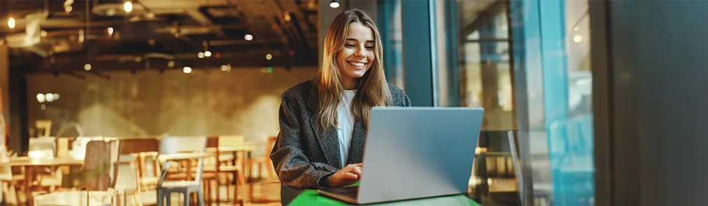 Uma jovem mulher sorridente trabalha em seu laptop em um café ou espaço de co-working moderno. A imagem sugere que ela está realizando a Gestão de contratos e documentos de forma digital e eficiente, aproveitando a flexibilidade do ambiente de trabalho.