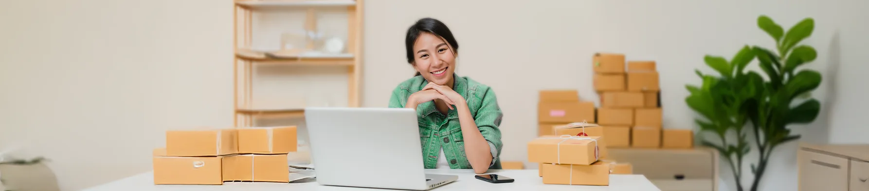 Uma mulher sorridente sentada em uma mesa de trabalho com seu laptop e pilhas de caixas de remessa ao redor, em um ambiente de escritório caseiro. A imagem representa o empreendedorismo e a eficiência digital, destacando a necessidade da assinatura eletrônica para pequenas empresas gerenciarem seus contratos de venda e logística.
