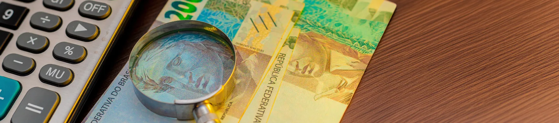 Calculadora, lupa e notas de Real sobre uma mesa de madeira, representando a organização e análise para a declaração do Imposto de Renda.