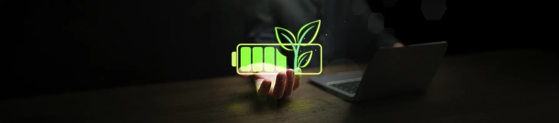 Mão exibe ícone luminoso de bateria com folhas ao lado de laptop, ilustrando a due diligence socioambiental.